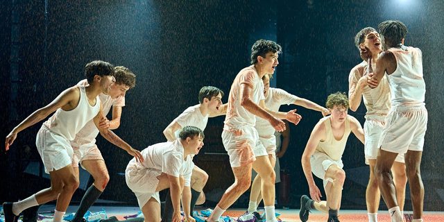 A group of boys in white vests and shorts are writhing around the stage which is being showered with blood. There are sweets and packets of crisps scattered across the floor.