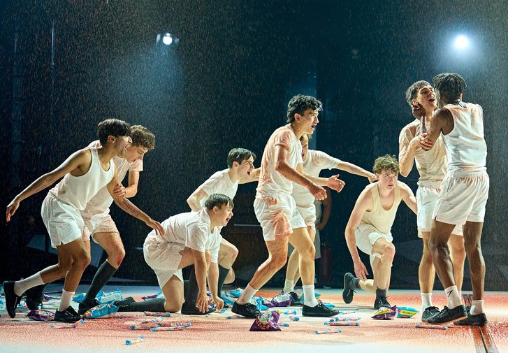 A group of boys in white vests and shorts are writhing around the stage which is being showered with blood. There are sweets and packets of crisps scattered across the floor.