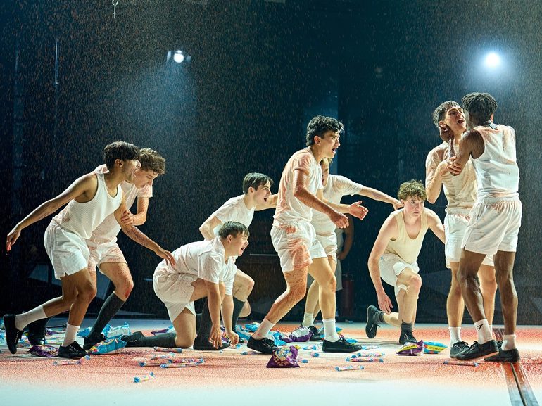 A group of boys in white vests and shorts are writhing around the stage which is being showered with blood. There are sweets and packets of crisps scattered across the floor.