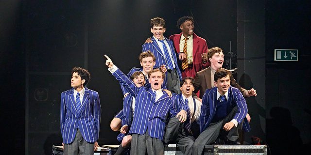 A group of boys dressed in school uniforms sit among several flight cases. They are all laughing and mucking around.