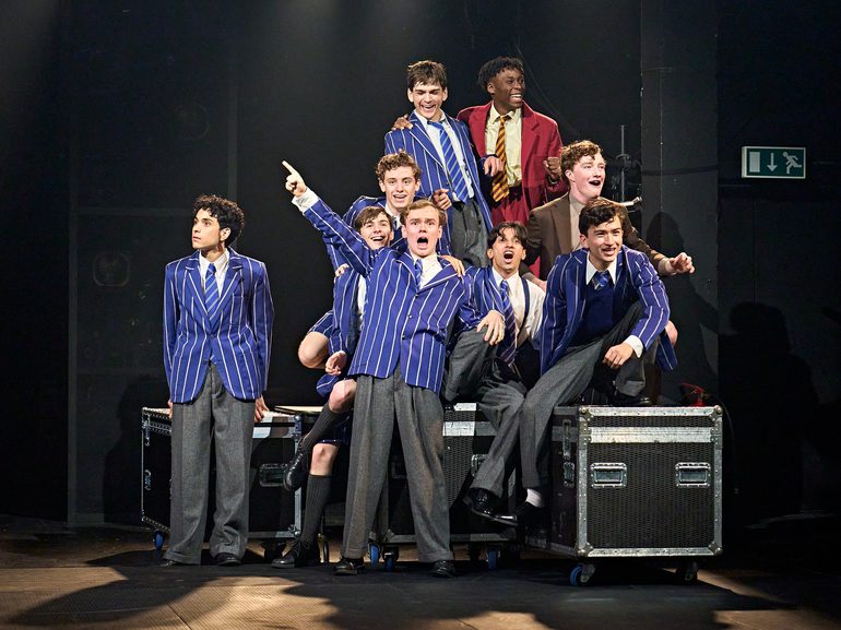 A group of boys dressed in school uniforms sit among several flight cases. They are all laughing and mucking around.