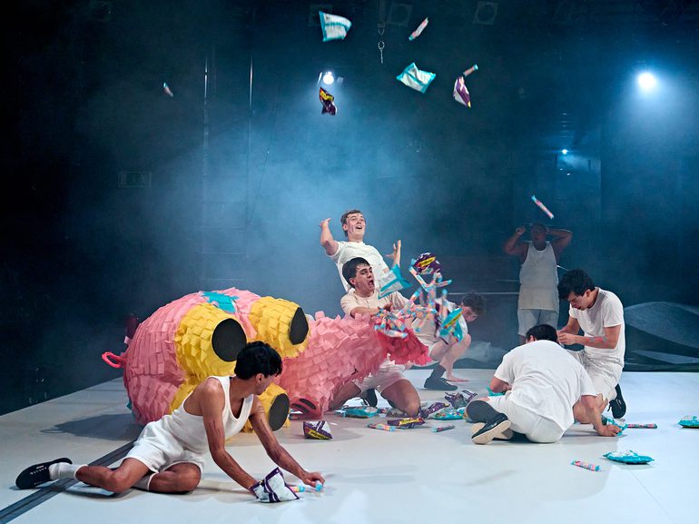 A group of boys in white vests and shorts are writhing around the stage. A large pig piñata is torn and there are sweets and packets of crisps spilling out, scattered across the floor.