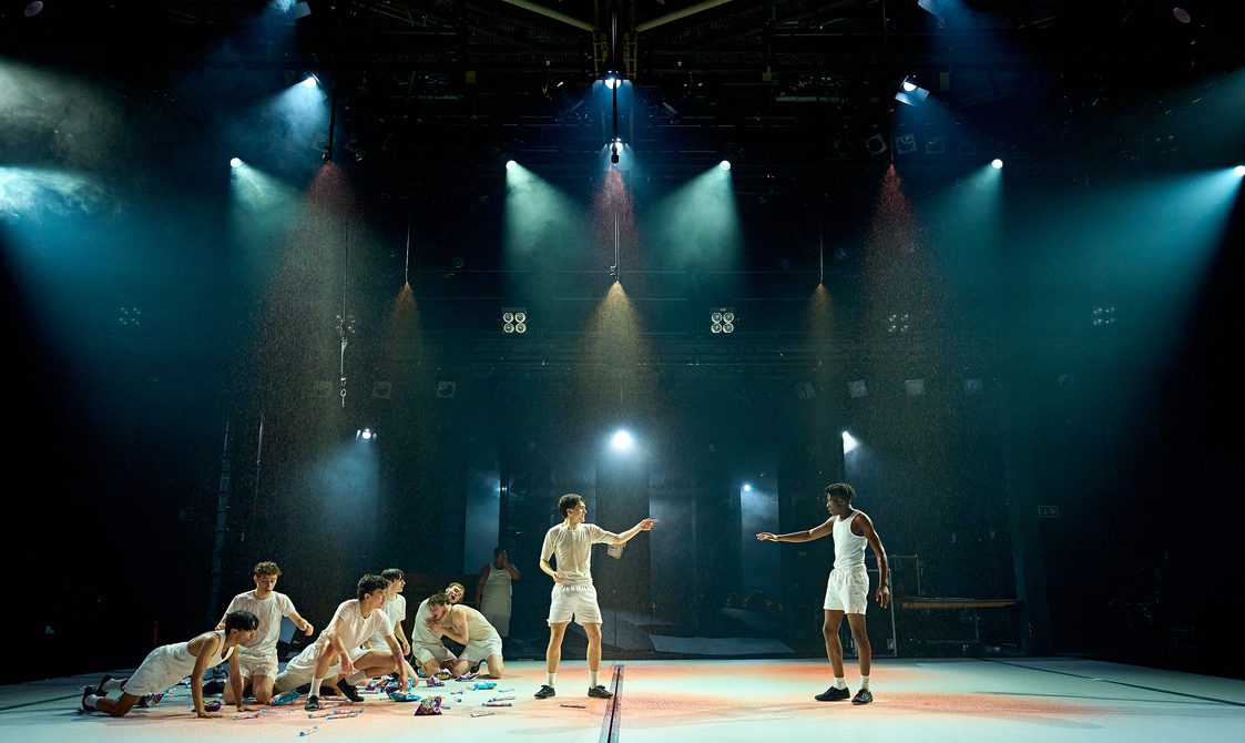 Two boys stand pointing at one another. To the left, a large group of boys are gathered, crouching down with sweets and packets of crisps scattered around them. Blood is raining down over the stage.