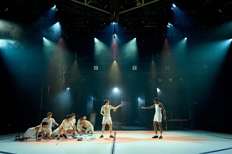 Two boys stand pointing at one another. To the left, a large group of boys are gathered, crouching down with sweets and packets of crisps scattered around them. Blood is raining down over the stage.