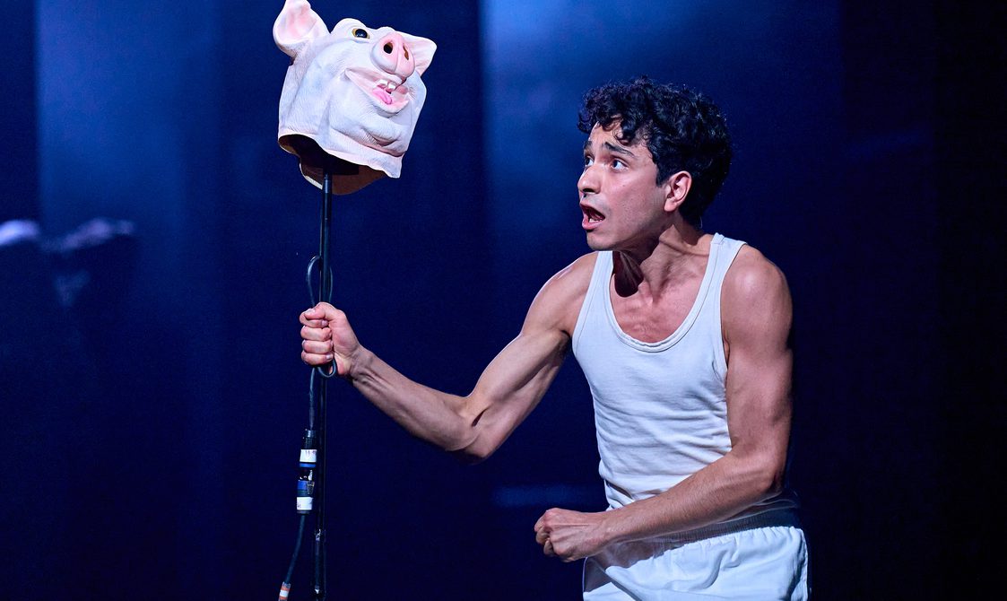 A boy in a white vest stares at a rubber pig mask which is on top of the microphone stand he is gripping tightly. He looks pained.