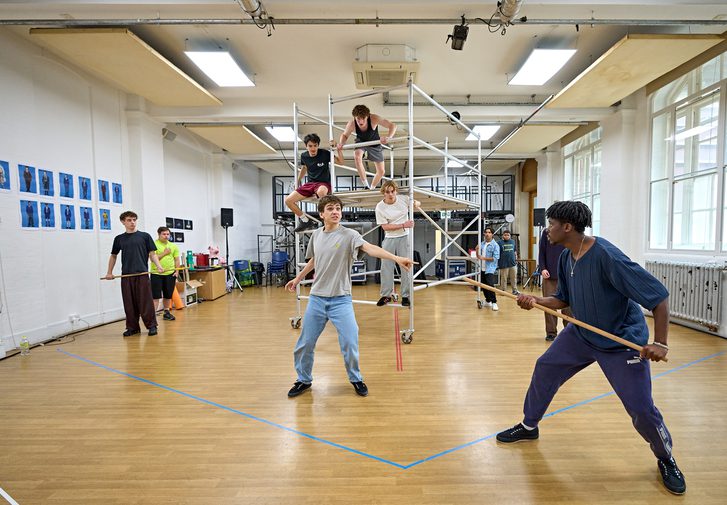 A group of performers are in a rehearsal room. There is a scaffolding rig in the middle of the room which several boys are climbing on. Others are stood around holding sticks. In the front and centre, one boy is pointing to the boy with a stick on the right. He looks panicked.