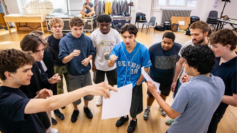 A large group of performers are gathered reading from a piece of paper being held up by the boy on the left. They have focussed expressions.
