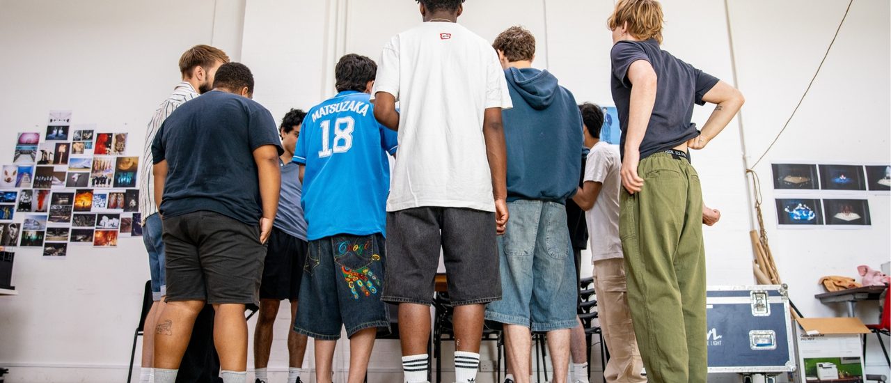 A group of boys are stood with their backs to the camera. They are all wearing casual clothes and are stood close together in a rehearsal room.