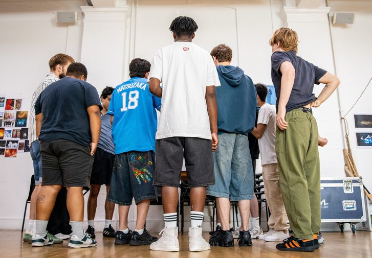 A group of boys are stood with their backs to the camera. They are all wearing casual clothes and are stood close together in a rehearsal room.