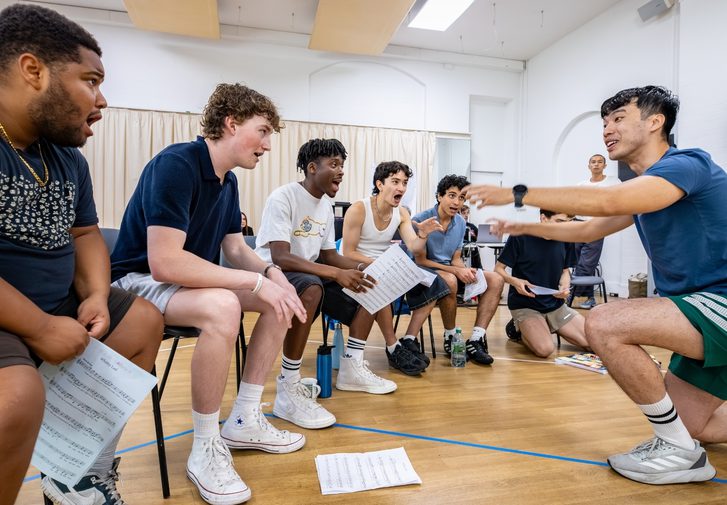 A group of boys are sat in a row singing intensely. They are all facing the musical director (Chris Poon) who is knelt in front of them conducting.