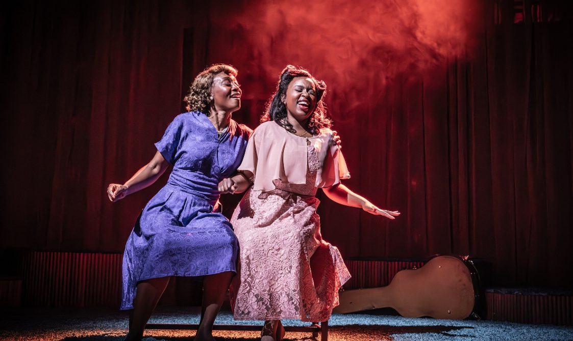 Two Black women are sat down. They both have their arms out as if dancing and have brilliant smiles on their faces. The lighting on the stage is red and there is a faint haze behind them.