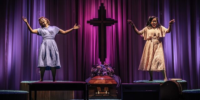 Two Black women are singing passionately with their eyes closed and arms extended freely on two raised platforms either side of a large crucifix with flowers at the base. There are luxurious indigo and magenta curtains flowing behind them.