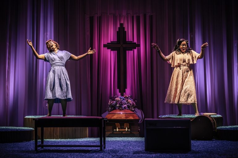 Two Black women are singing passionately with their eyes closed and arms extended freely on two raised platforms either side of a large crucifix with flowers at the base. There are luxurious indigo and magenta curtains flowing behind them.
