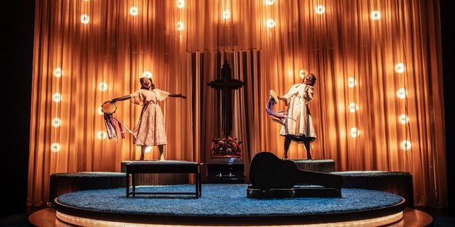 Two Black women are singing passionately on two raised platforms either side of a large crucifix with flowers at the base. They are both holding tambourine that have purple and white ribbons flowing from them. The backdrop has warm marquee style lighting with dramatic curtains. There is a piano stool and guitar case at the front of the stage.