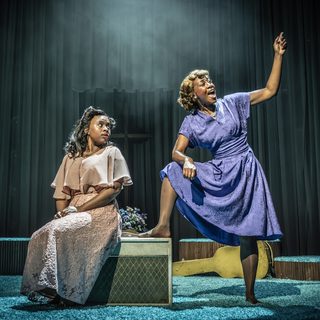 Two Black women are in the centre of a stage covered with teal shag carpet. The younger woman on the left is sat on a box with her hands in her lap, looking over her shoulder at the older woman on the right who is confidently stood with one foot propped up on the box. She is singing powerfully, facing away from the other woman with one arm raised.