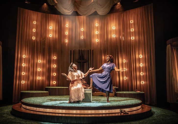 Two Black women are singing joyfully with their arms extended expressively facing each other. The younger woman on the left is sat on a box wearing a pale pink floral dress with a flowing shawl and the older woman on the right who is confidently stood with one foot propped up on the box. She is barefoot and wearing a muted blue knee length dress. Their styling is evocative of the 1940s.