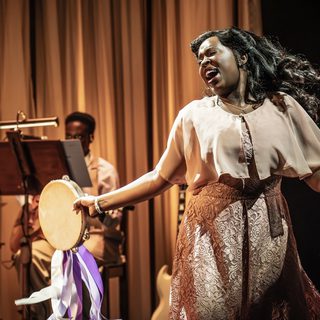 A young Black woman is singing passionately with a tambourine that has cascading purple and white ribbons in her hand. Her hair is in 1940s style victory rolls, with a flowing pale pink shawl over a floral jacquard dress in the same colour and a simple crucifix around her neck. There is a musician playing the guitar in the background with a music stand in front of them.