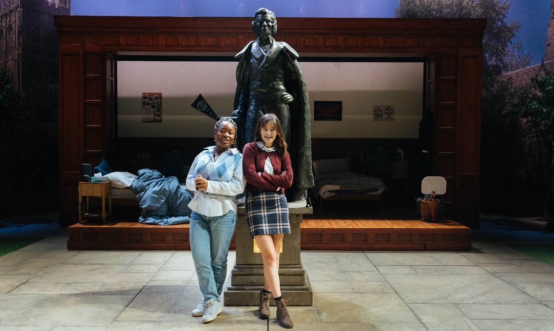 Two young women are stood in front of a large bronze statue of a man. They both stand with their arms crossed and look mischievous. There is a messy dorm room in shadow in the background.