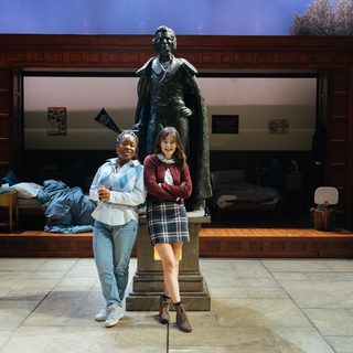 Two young women are stood in front of a large bronze statue of a man. They both stand with their arms crossed and look mischievous. There is a messy dorm room in shadow in the background.