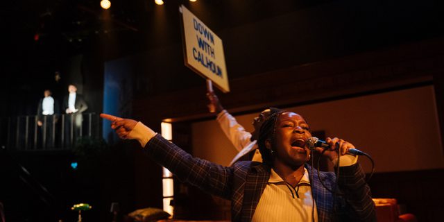 A woman wearing a tartan blazer is shouting passionately into a microphone. Her arm is stretched out to her side and she is pointing with her index finger. Behind her, a man is also shouting passionately and is holding aloft a placard that reads 'Down with Calhoun' in bold capital letters.