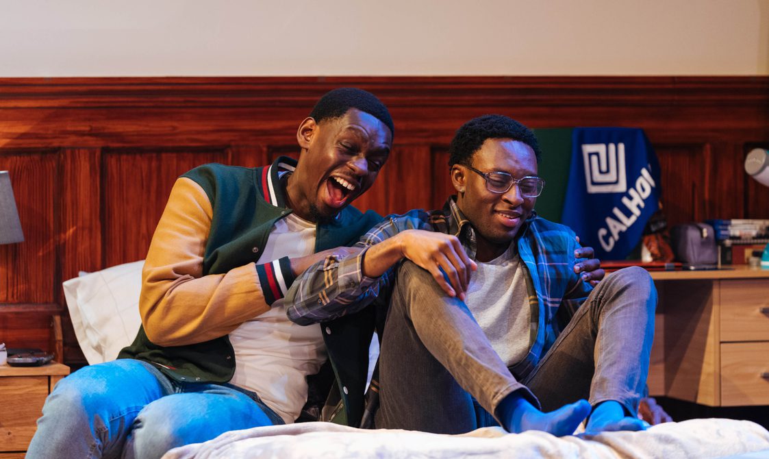 Two young men are sat on a bed. One is laughing hysterically and grabbing the other's arms, while the other smiles awkwardly.