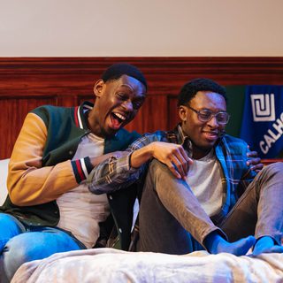 Two young men are sat on a bed. One is laughing hysterically and grabbing the other's arms, while the other smiles awkwardly.