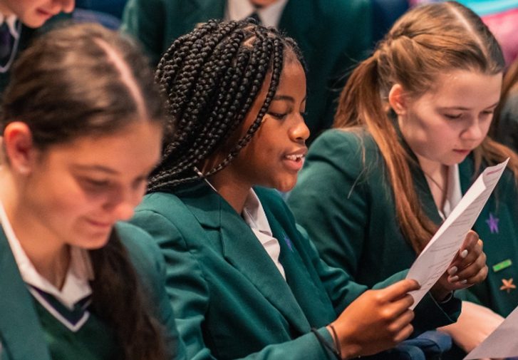 A groups of school children in green blazers and white shirts sit reading scripts in the theatre.