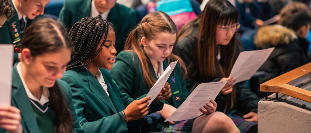 A group of school children in green blazers and white shirts sit reading scripts in the theatre.