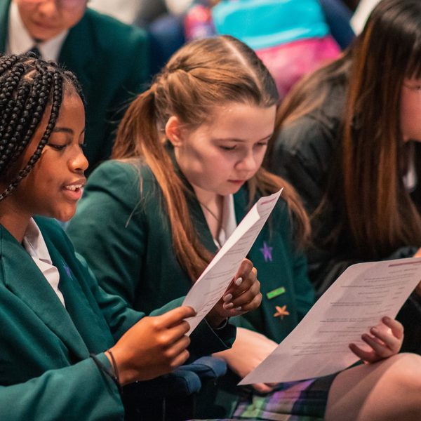 A group of school children in green blazers and white shirts sit reading scripts in the theatre.