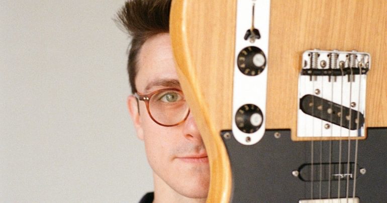 A close-up of a man with dark hair and glasses who is smirking. Half of his face is obscured by a wooden electric guitar being held up in front of him.