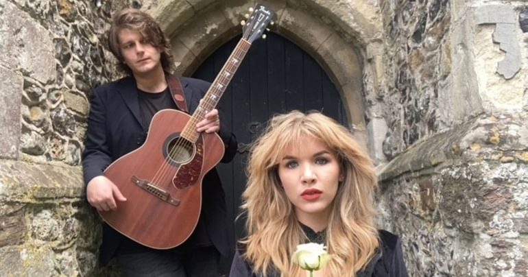 Two people are in a doorway of an old stone building. On the left, a man with light brown hair, dressed in black, is holding an acoustic guitar. He is looking down at the woman on the right of the picture, who is looking directly at the camera. She has voluminous blonde hair and is wearing a black top and red lipstick. She is holding a white rose.