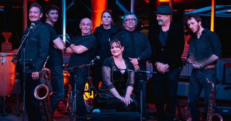A group of eight people all wearing black and smiling. Seven are stood posing at the camera. In front, there is a woman sat down with her hands in her lap. There are two saxophones and three microphones.