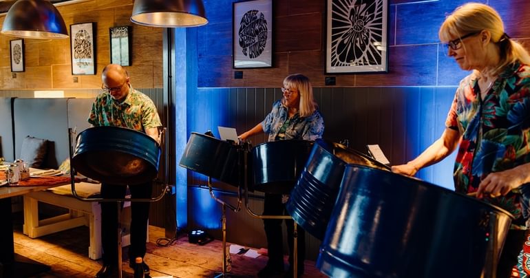 Three people are stood playing steel drums. They all have glasses and are wearing colourful Hawaiian shirts. There is deep blue lighting behind them.