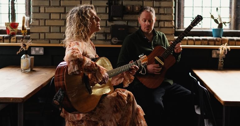 Two people are sat in front of a brick wall with potted plants dotted around. They are both playing acoustic guitars. On the left, a woman with blonde curly hair is wearing a flowy floral dress. She is singing. On the right, a man with dark hair and facial hair is wearing a dark green shirt and dark trousers.