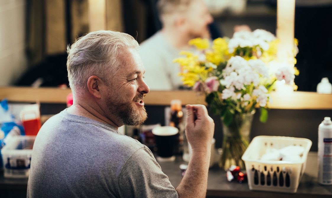 An actor sat at a dressing room table, he is smiling as he talks to someone out of shot.