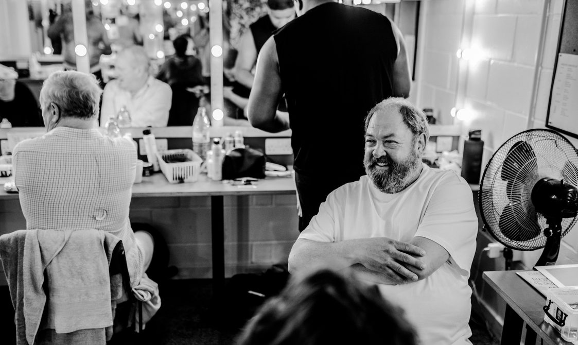 An image of a dressing room before a show - in the background two actors are looking in the mirror, in the foreground Mark Addy is sat smiling with his arms crossed.
