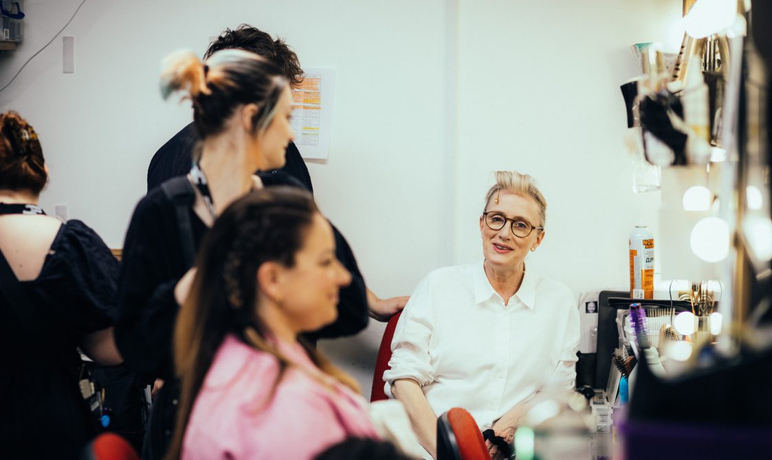 In the foreground, an actor wearing a pink shirt is having a hair braided, in the background an acotr wearing a white shirt sits smiling at her.