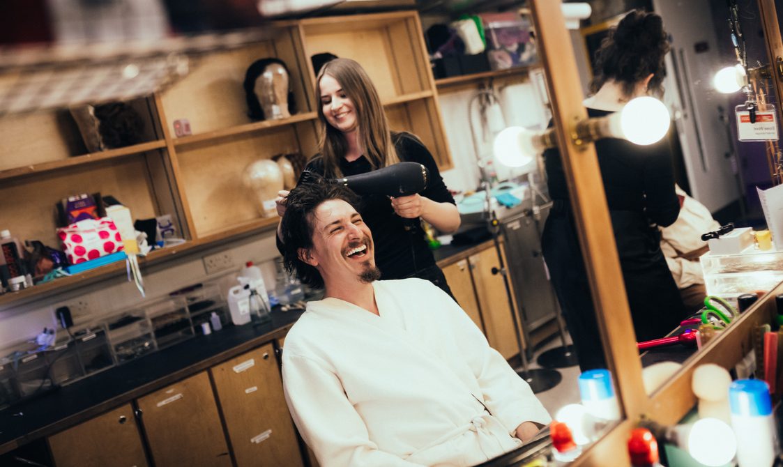 Tom Rosenthal sat at a dressing room table having his hair blow dried - he is laughing and wearing a white robe.
