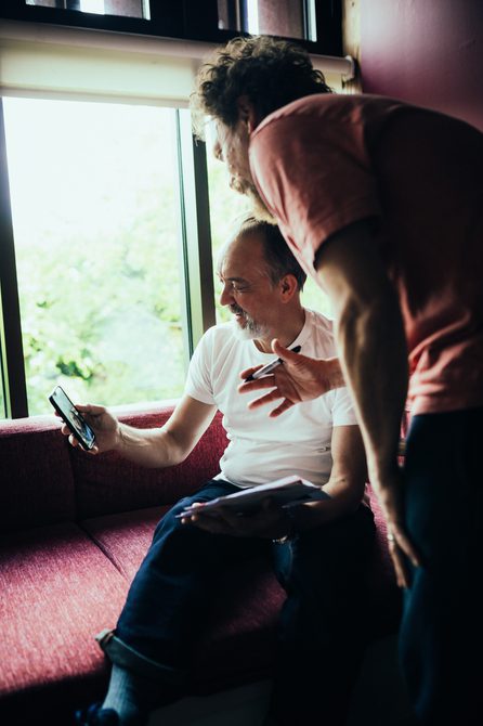 An actor is sat in a dressing room showing another actor something on his phone - they're both looking at the phone with an interested expression.