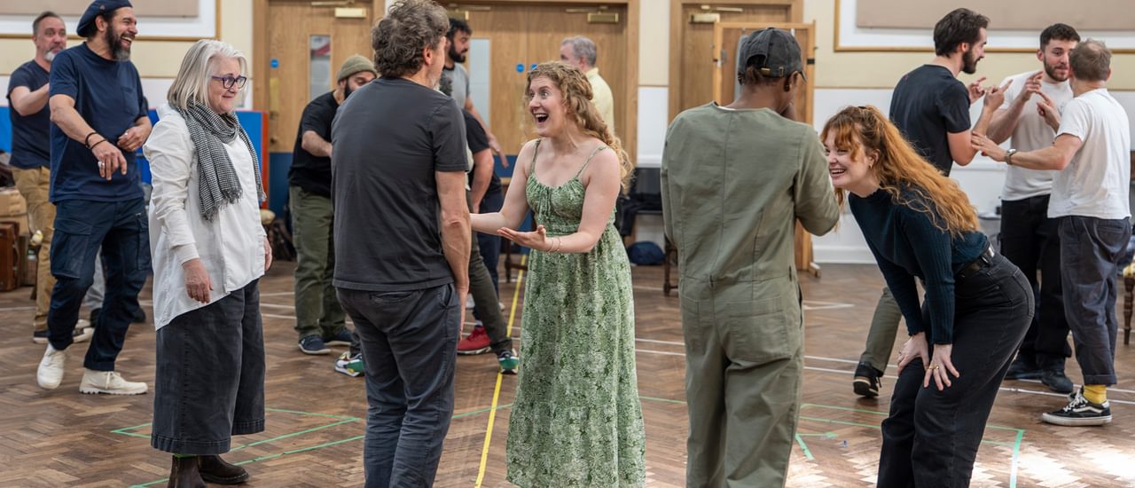 Performers wearing casual clothes rehearse in a studio. The room is crowded with smaller groups forming, talking and laughing together. In the centre, four performers stand facing one another in two lines. The two women facing the camera are laughing and smiling with animated expressions.