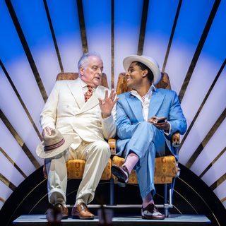 Two men are sat talking to one another. On the left, a man in a cream suit is resting a straw fedora on his knee. On the right, a man in a powder blue suit and a cream fedora is smiling widely at the other man.