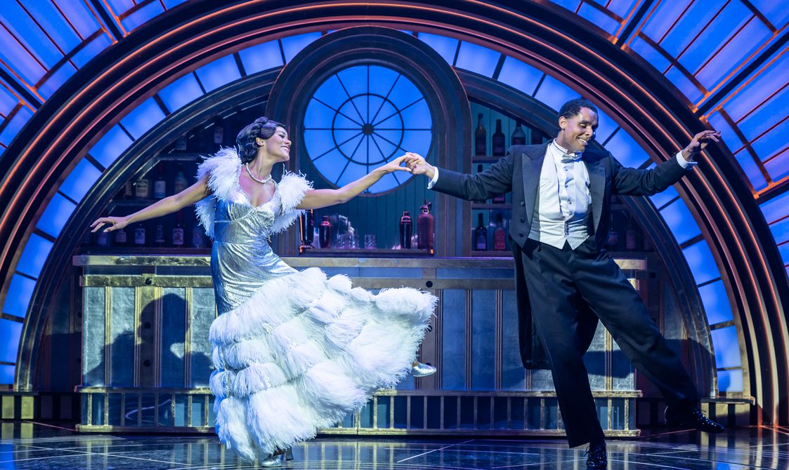 A man and woman are dancing elegantly side by side while holding hands. She is wearing a exquisite silver gown with a white ostrich feather skirt and detailing on the sleeves. He is wearing white tie and tails. They are smiling sweetly. Behind them is a glamourous blue and gold 1930s art deco bar.