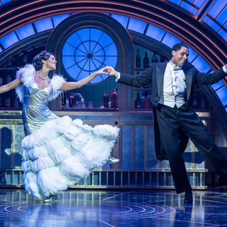 A man and woman are dancing elegantly side by side while holding hands. She is wearing a exquisite silver gown with a white ostrich feather skirt and detailing on the sleeves. He is wearing white tie and tails. They are smiling sweetly. Behind them is a glamourous blue and gold 1930s art deco bar.