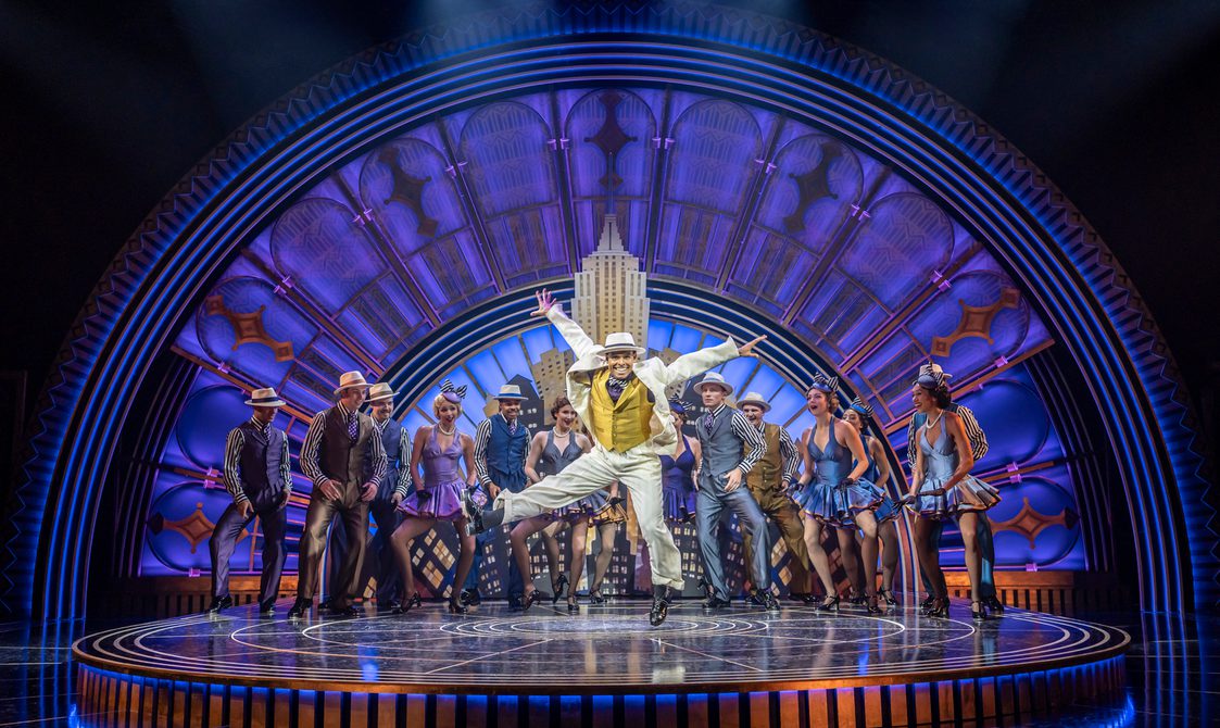 A company of dancers are all wearing refined 1930s outfits. The men have cream fedoras, matching waistcoats and trousers, striped shirts and polka ties. The women have mini A-line dresses and fascinators with striped bows. Front and centre, a man in a cream suit and matching hat with a mustard waistcoats dancing with his arms above his head and has legs kicking. The set is a detailed art deco arch with skyscrapers in front.