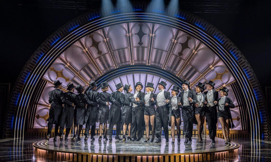 A large group of dancers are all wearing black top hats, black tails and white waistcoats with canes under their arms. They each have an arm around the waist of the person next to them with have facing the camera and the other half facing the back of the stage which has a black, gold and deep blue art deco arch.