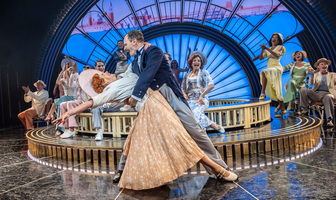 A man and woman are dancing. He is holding her by her waist as he dips her. She is leaning deeply with one arm reaching behind her and the other grasping his neck. He has on an sophisticated navy and grey tailed suit. She is wearing a light orange pleated skirt with white floral details, a white cropped jacket and large white hat. There are many performers behind them clapping and cheering. The backdrop is a large art deco arch with a projection of Venice.