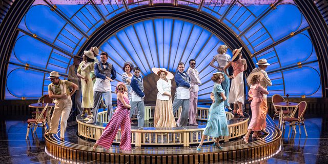 A group of performers all wearing elegant 1930s outfits. They are dancing with one hand wrapped around their front and the other behind their back. A large art deco arch has a projection of Venice.
