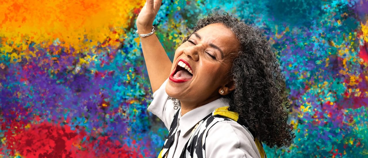 A woman sings joyfully, eyes closed and mouth open wide. Her head is passionately thrown back and her arm is raised. She has black and grey curly hair and wears a white short sleeved shirt with a black, yellow and white patterned waistcoat and bright red lipstick. The background is a bright array of colourful, dappled paint.