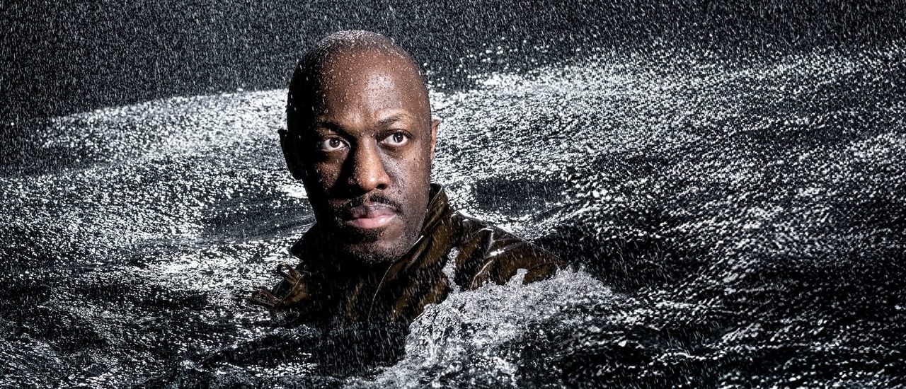 A man's head and shoulders rise from a sea of rough, churning water; rain is pouring down on him. He stares into the distance, his face illuminated by a shaft of light. He has dark hair and a moustache.