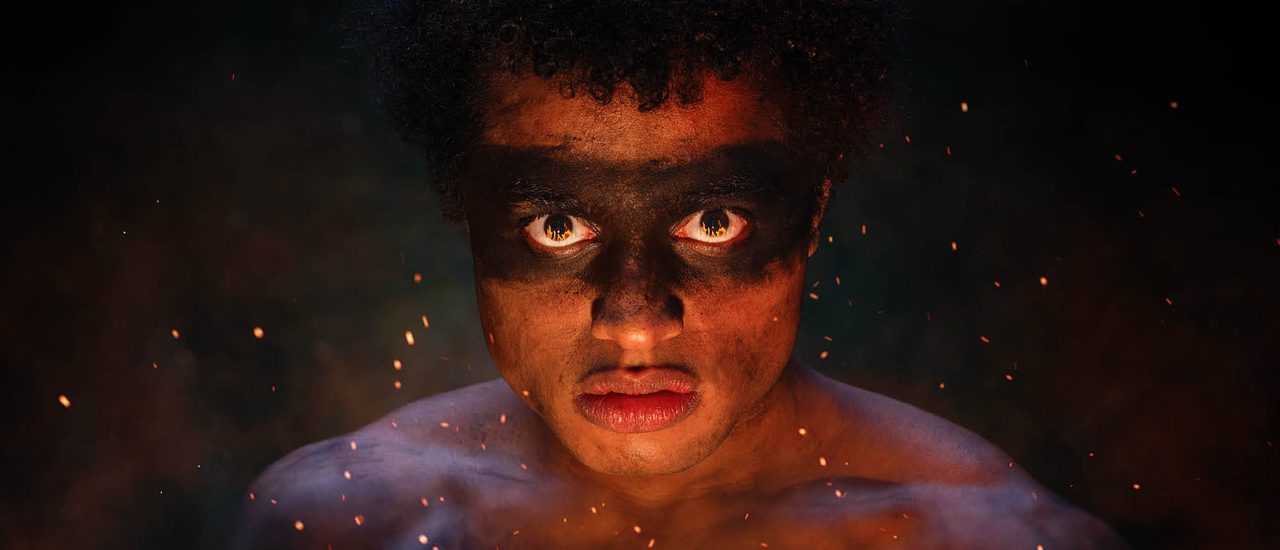 A young man is looking directly into the camera, with a serious look on his face. Flames are reflected in his eyes and sparks surround him.